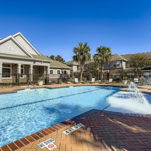 A view of a swimming pool area featuring a clear blue pool, brick paving, a splash fountain, and palm trees. There is a residential building in the background and a fence surrounding the pool.
