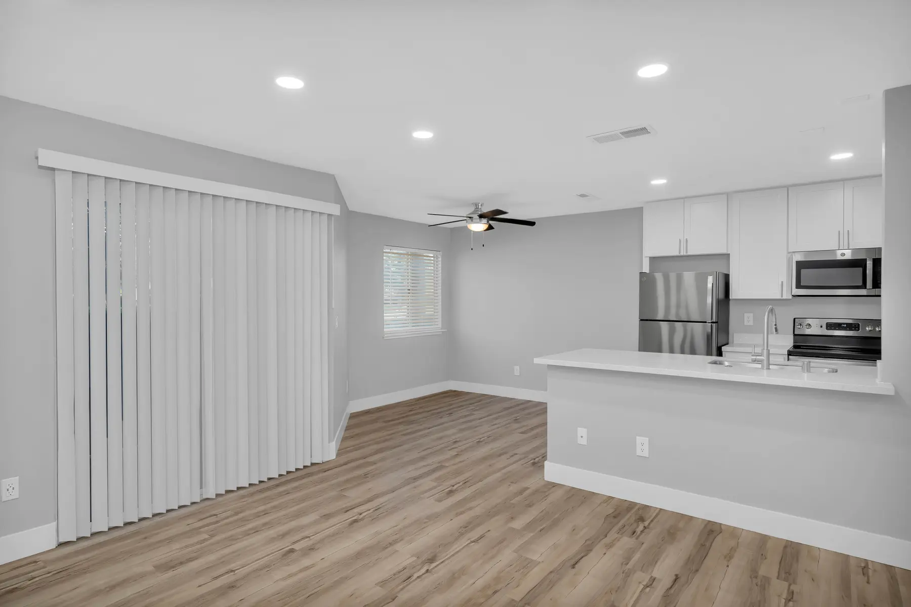 A modern open-concept living area featuring light gray walls, a ceiling fan, and white cabinetry. The kitchen includes stainless steel appliances, and there is a sliding glass door with vertical blinds leading to an outdoor space. The floor is covered with light wooden planks.