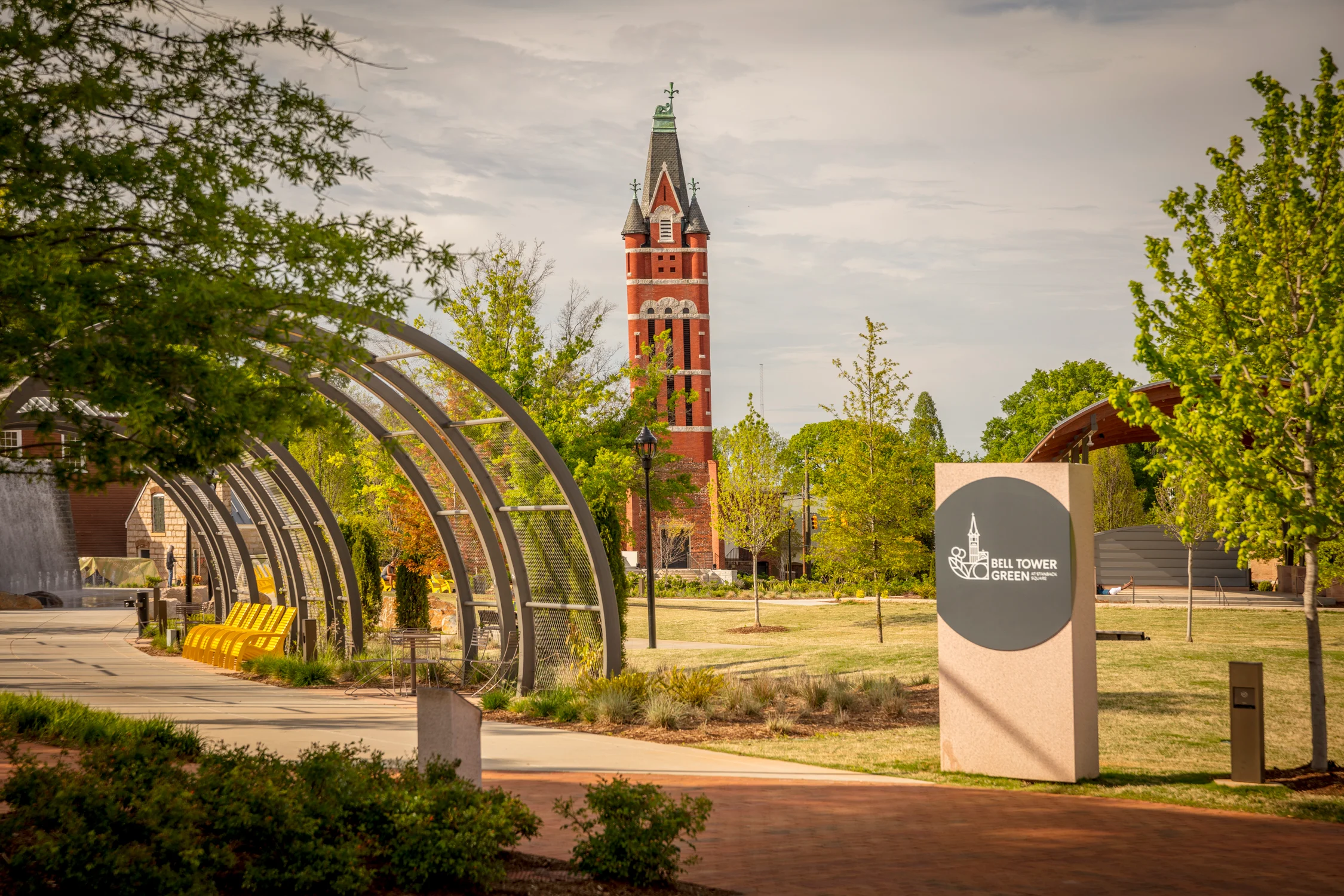 A park scene featuring a tall clock tower in the background, surrounded by green trees and a modern art structure in the foreground.