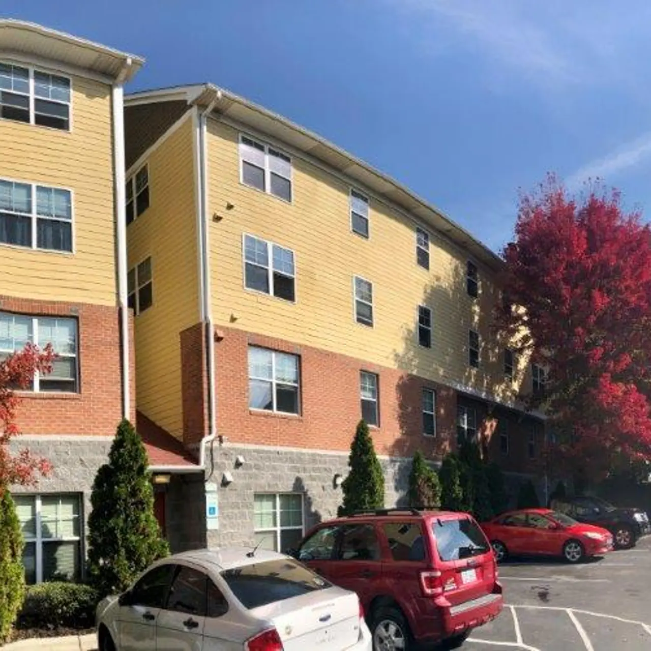 A multi-story apartment complex featuring a mix of yellow and red brick exteriors, surrounded by trees with autumn foliage. Several parked cars are visible in the foreground.