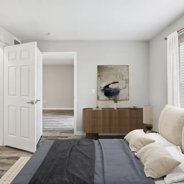 A modern bedroom featuring a neutral color scheme with a large bed, wooden dresser, and an open closet area. There is an abstract artwork on the wall and natural light streaming in from the window with blinds.
