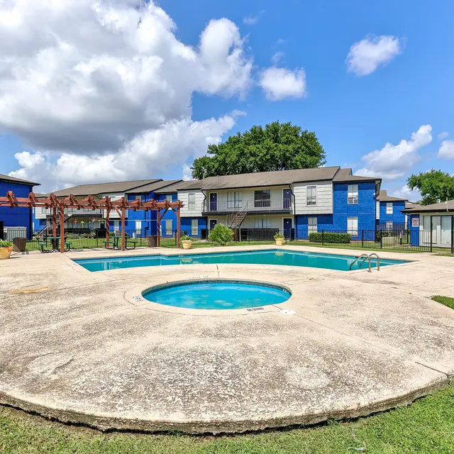 A swimming pool area in an apartment complex surrounded by blue buildings and green grass under a bright blue sky with fluffy white clouds.