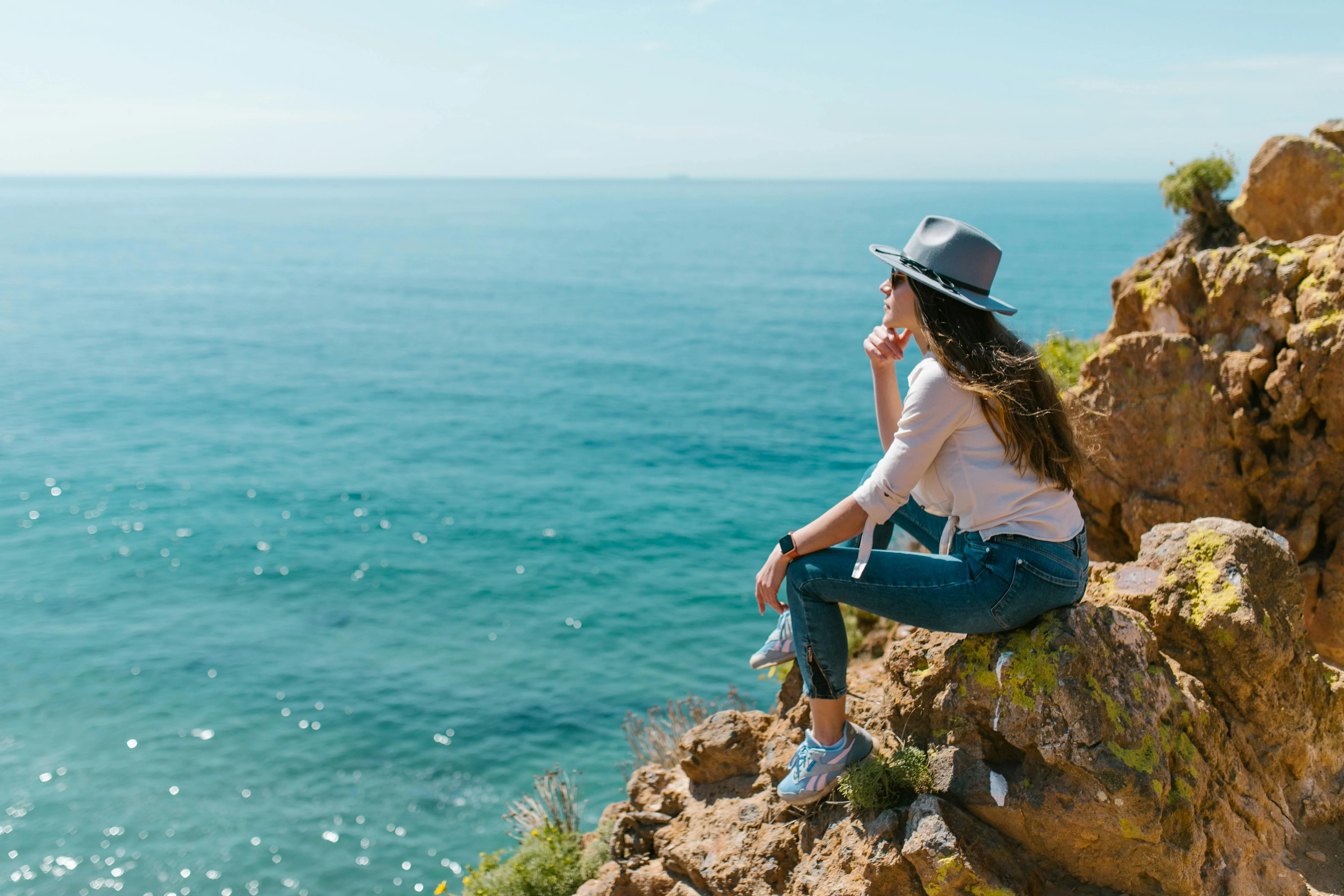 A Woman Contemplating by the Ocean A woman sitting on a rocky ledge overlooking the ocean, wearing a hat and casual attire, with her thoughtful pose reflecting tranquility.