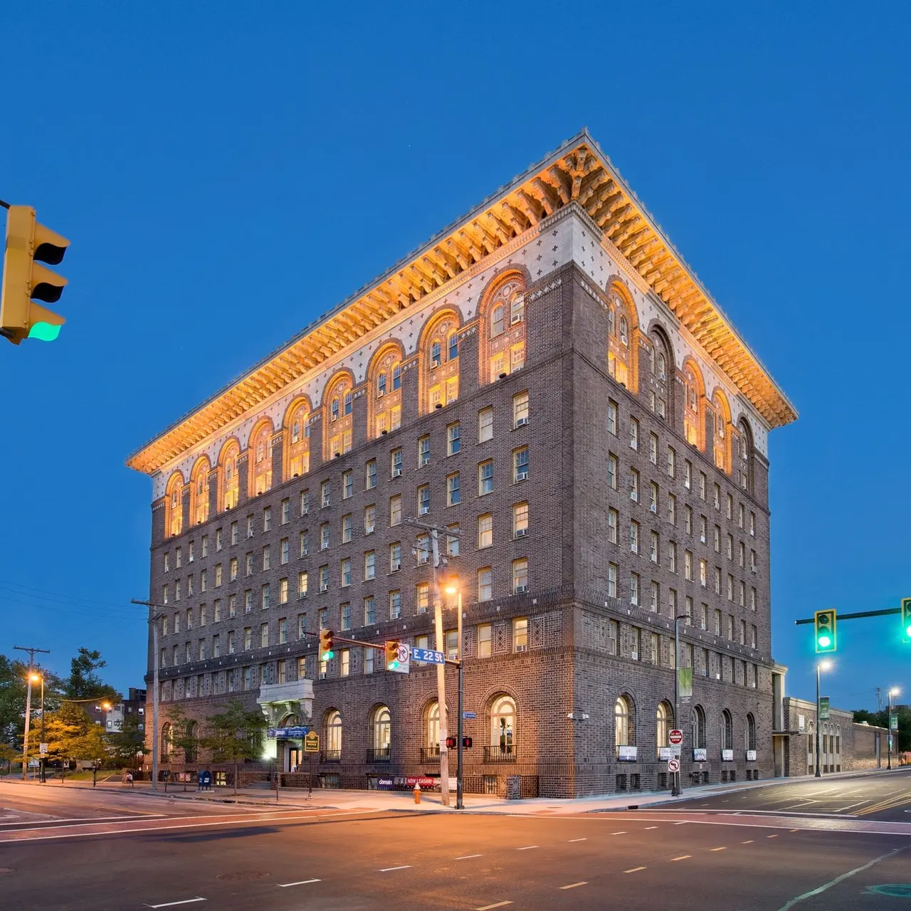 A historic brick building with illuminated windows, standing on a street corner during twilight. Traffic lights are visible, and the sky is a deep blue.