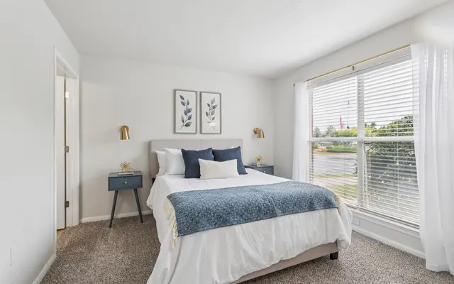 Bright bedroom with a queen bed, blue accents, two bedside tables, and large windows with sheer curtains.