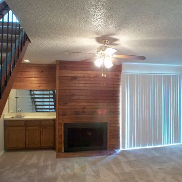 A cozy living room featuring a wooden accent wall, ceiling fan, and a fireplace, with large windows covered by vertical blinds. A staircase is visible in the background, leading to an upper level.