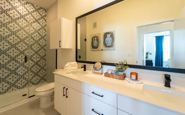 A modern bathroom featuring a large mirror, white cabinetry, and a stylish patterned wall. There is a shower area with glass doors and a large sink counter with decorative items.