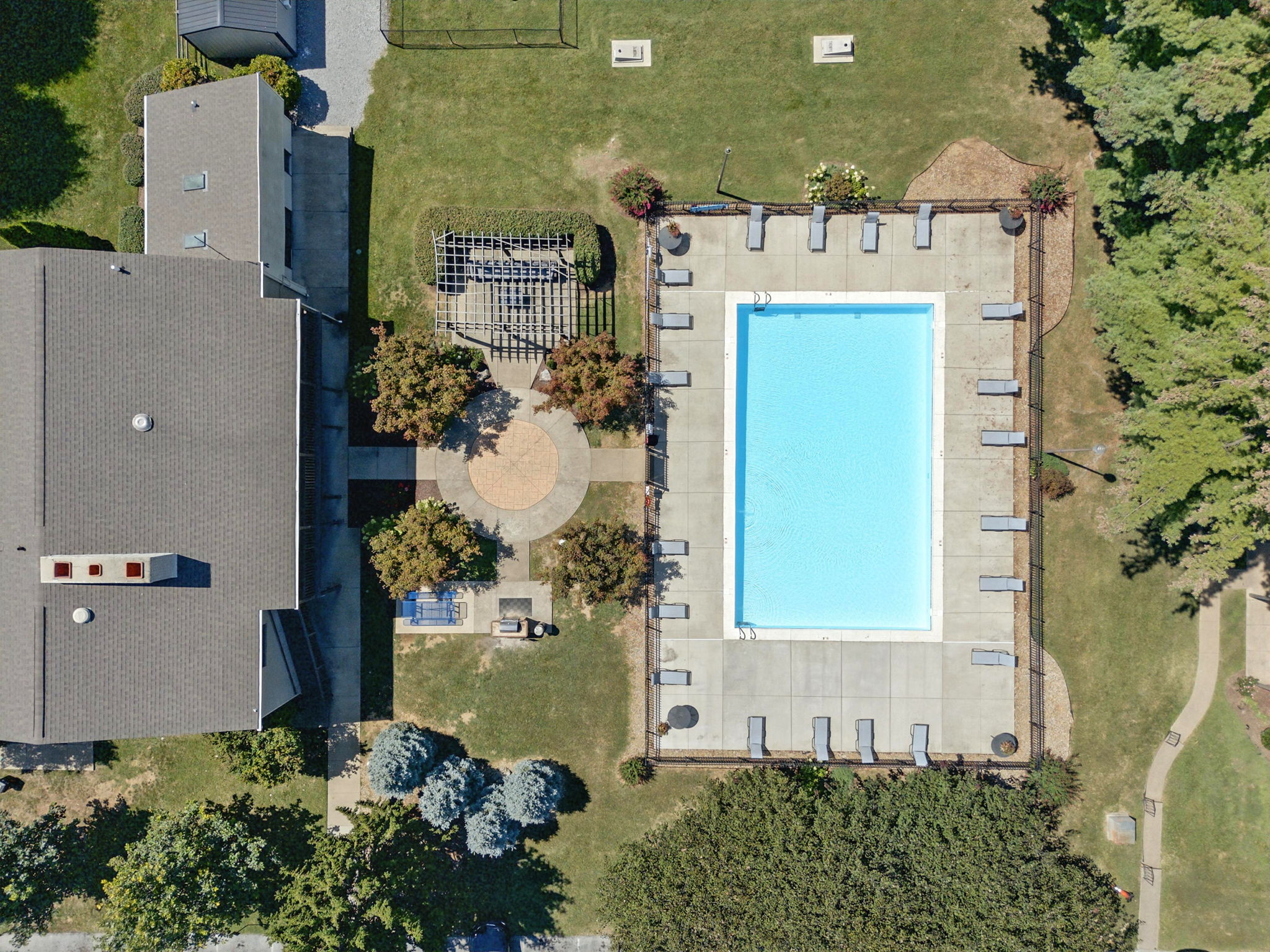 Aerial View of Pool Area Aerial view of a swimming pool surrounded by a concrete deck, lounge chairs, and landscaped greenery, with a housing structure visible nearby.