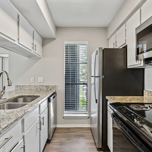 A modern kitchen featuring granite countertops, stainless steel appliances, and white cabinetry. The kitchen includes a double sink, a refrigerator, and an oven with a stove top.