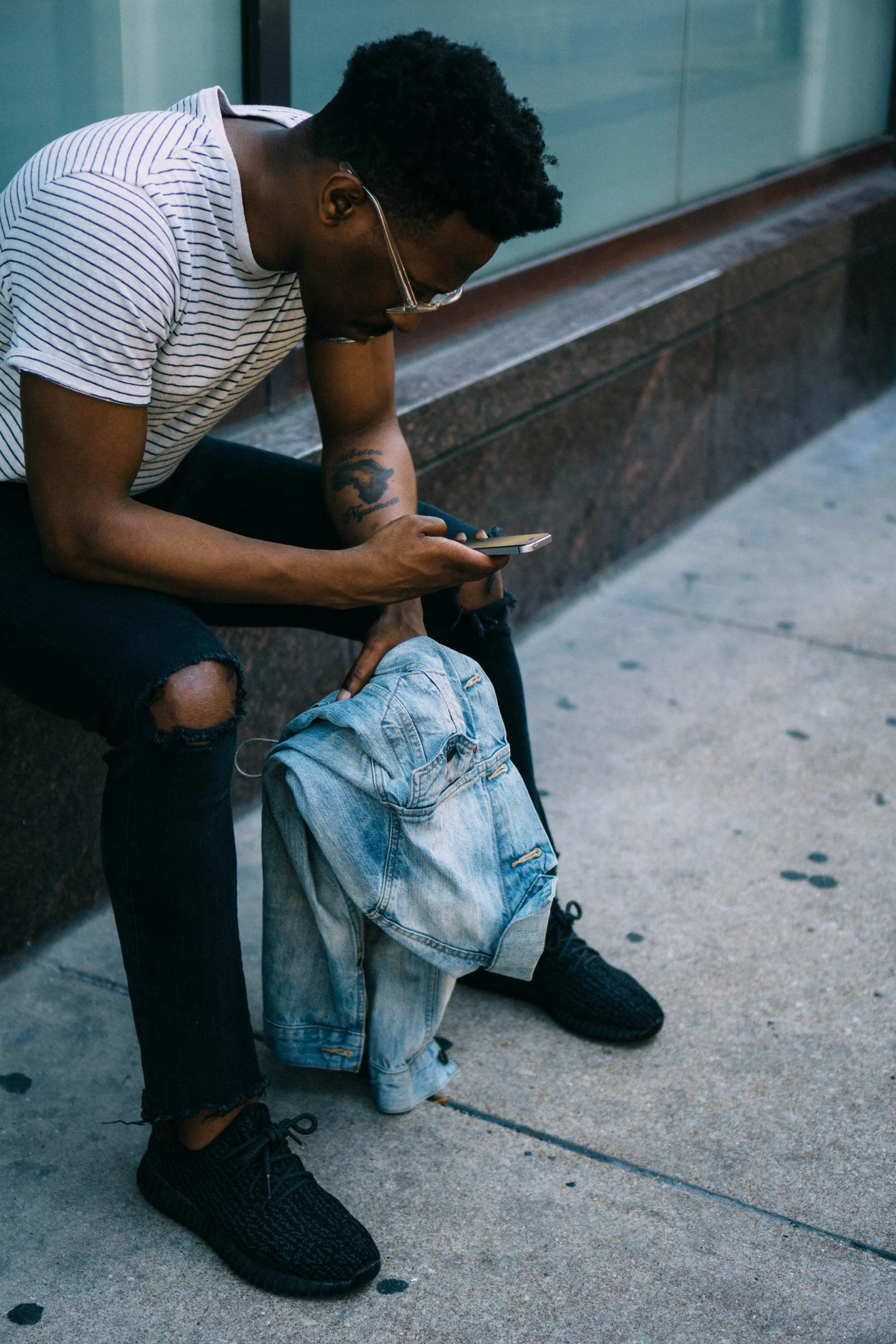A person sitting on a sidewalk, wearing a striped shirt and black jeans, looking down at their phone. They have a denim jacket draped over one arm and are wearing black shoes.