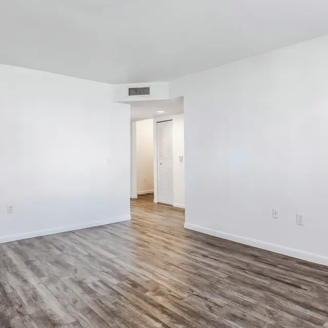 An empty room with light-colored walls and wood-like flooring, featuring a door to the left leading to another room.