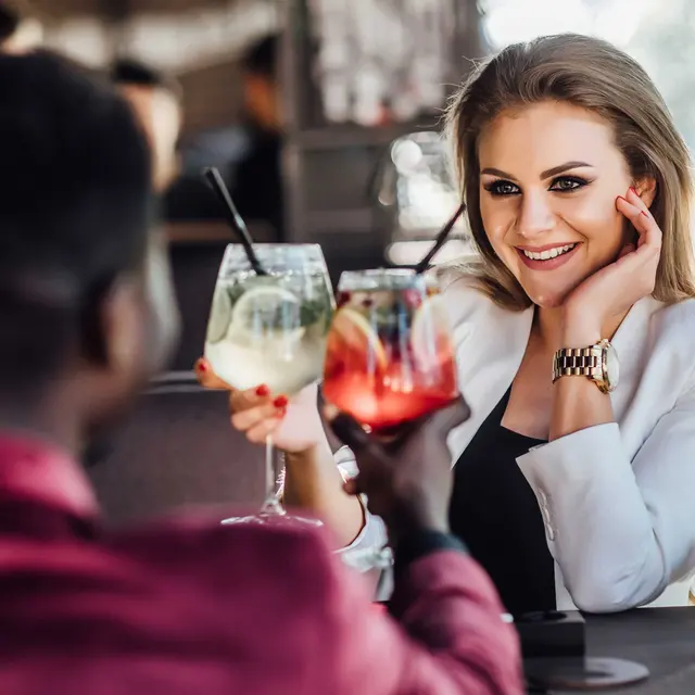 A couple sitting at a cafe table, toasting with colorful drinks. The woman is wearing a white blazer and smiling, while the man is dressed in a pink blazer. The background shows a blurred cafe setting with soft lighting.