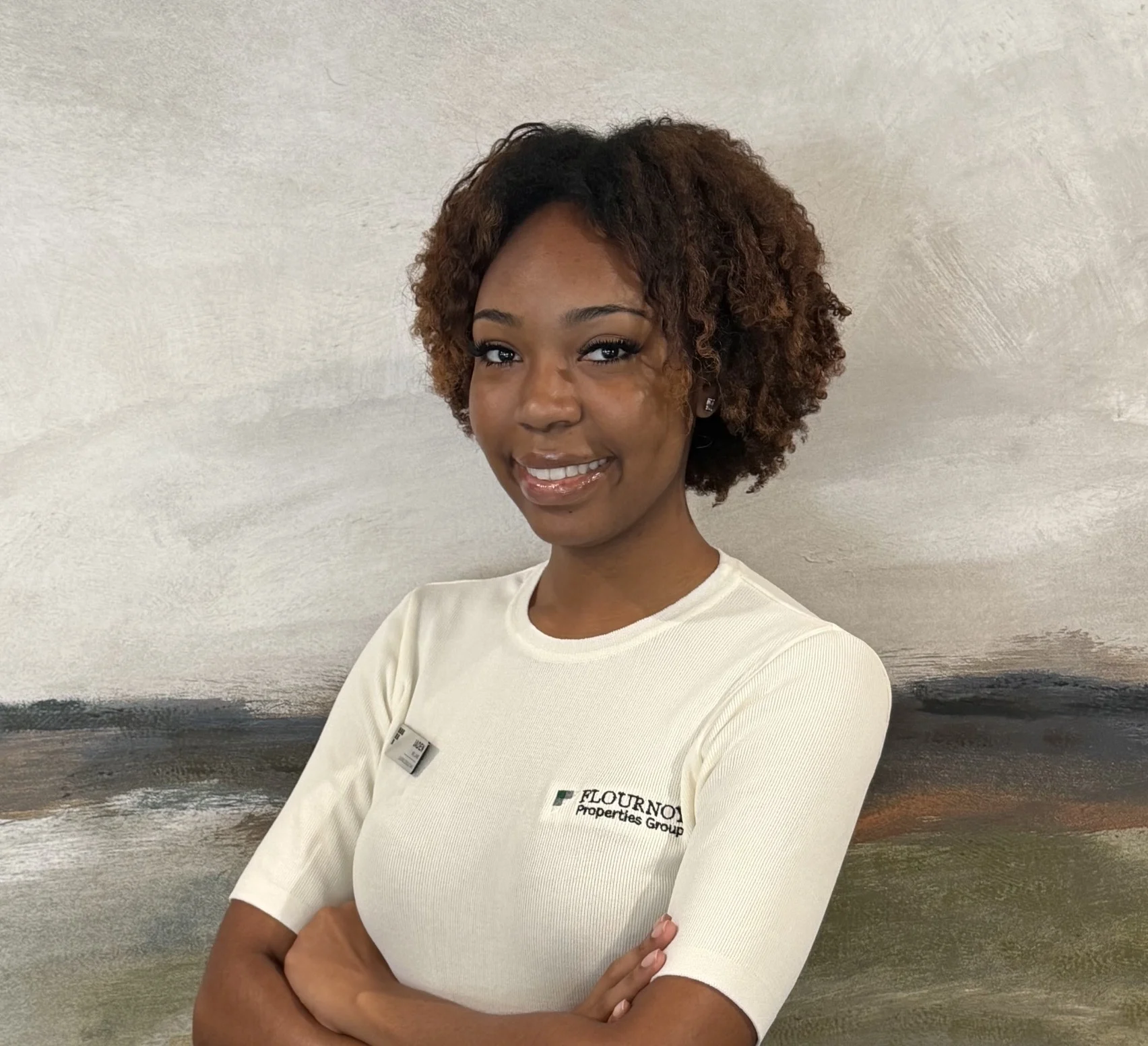 Professional Portrait of a Young Woman A smiling young woman in a white shirt with short sleeves, standing with arms crossed against a soft, abstract background.