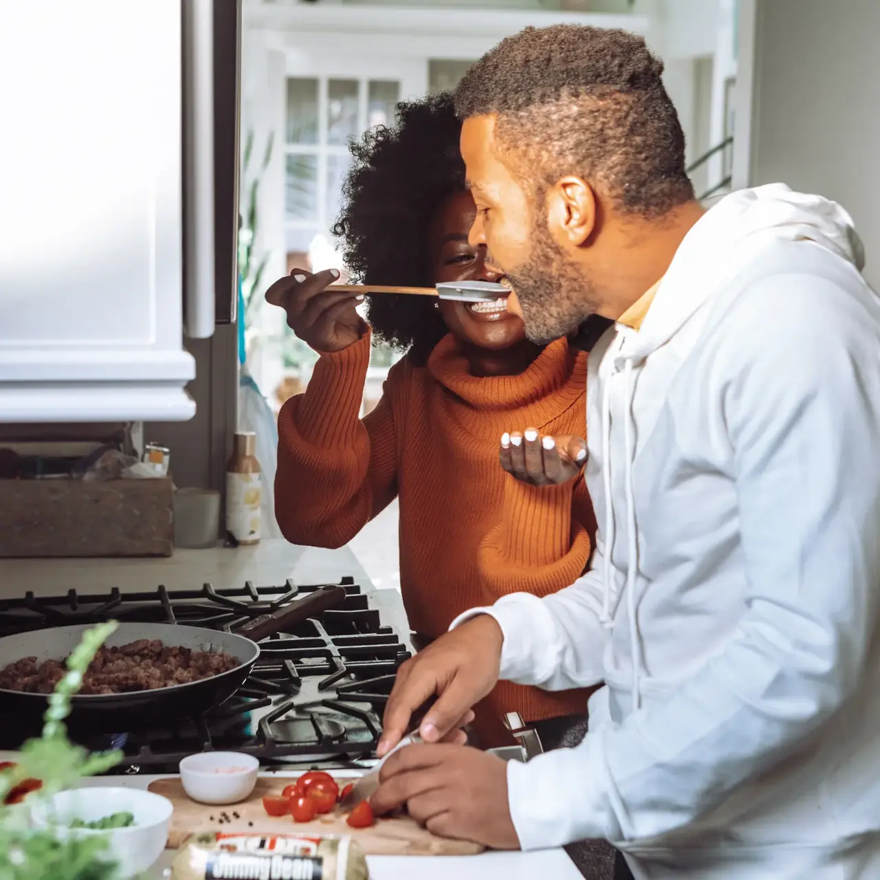 A couple cooking in a kitchen, with one person feeding the other. The woman in an orange sweater playfully holds a spoon, while the man in a white hoodie is chopping vegetables.