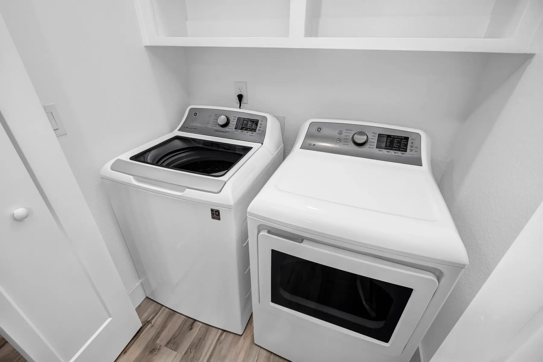 A modern laundry room with a washing machine and a dryer side by side, both in white. The space features light-colored walls and wooden flooring.