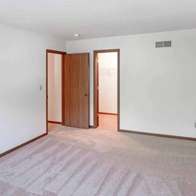 Court Place - Bedroom An empty bedroom with beige carpeting and white walls. There's a closed brown door on the left, a slightly open brown door in the center, and another open door on the right that leads to a closet.