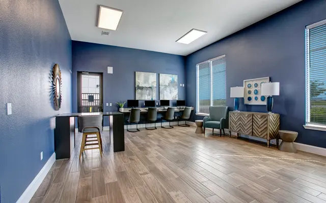 A modern office space featuring blue walls, wooden flooring, and ample natural light. It includes a long table with chairs, a seating area with a patterned chair, and decorative elements on the walls.