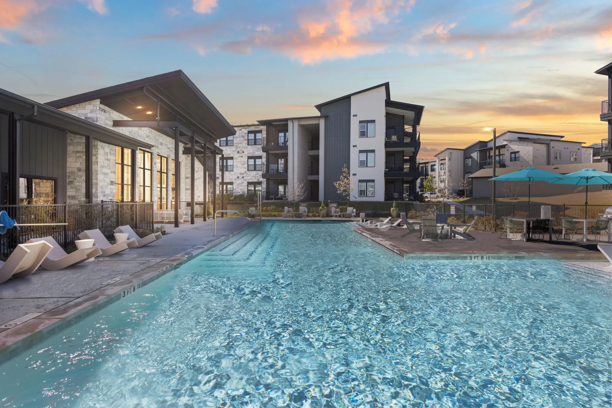View of a modern apartment complex with a swimming pool and lounge area during sunset.