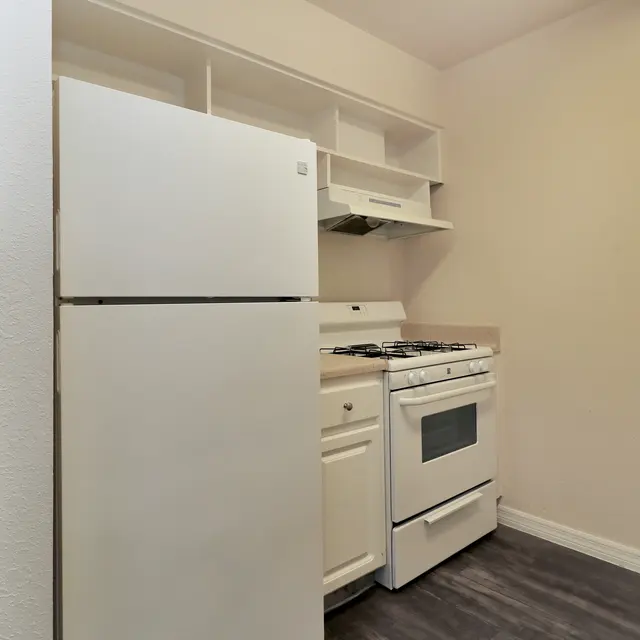 A small kitchenette featuring a white refrigerator and a white gas stove with an overhead range hood. The space has light-colored walls and a portion of the floor is visible, showing dark laminate flooring.