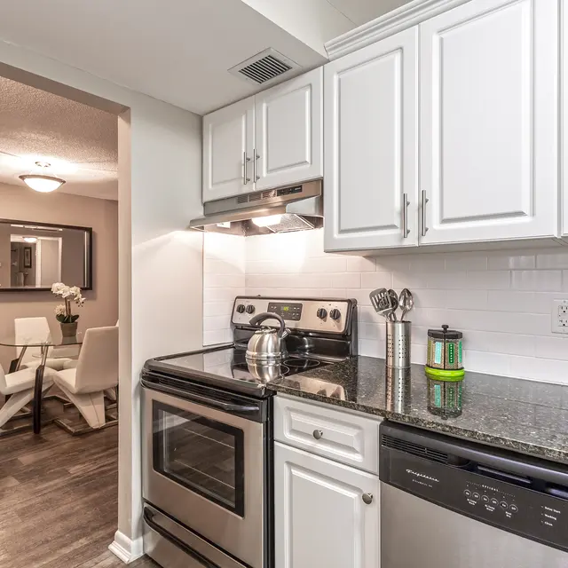 Kitchen 2 - Biscayne Apartments A modern kitchen featuring stainless steel appliances, including an oven and a dishwasher, alongside granite countertops and white cabinetry. A doorway leads to a dining area with a table and chairs in the background.