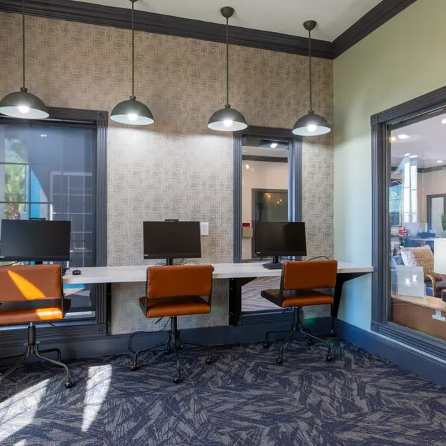 A modern office space featuring a shared desk area with three chairs and computers, illuminated by pendant lights, with a cozy lounge area visible through a window.