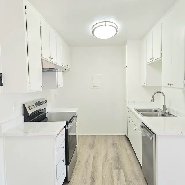 A bright and clean kitchen with white cabinets, stainless steel appliances, and modern fixtures. The floor is light-colored wood, and there is a circular ceiling light providing illumination.