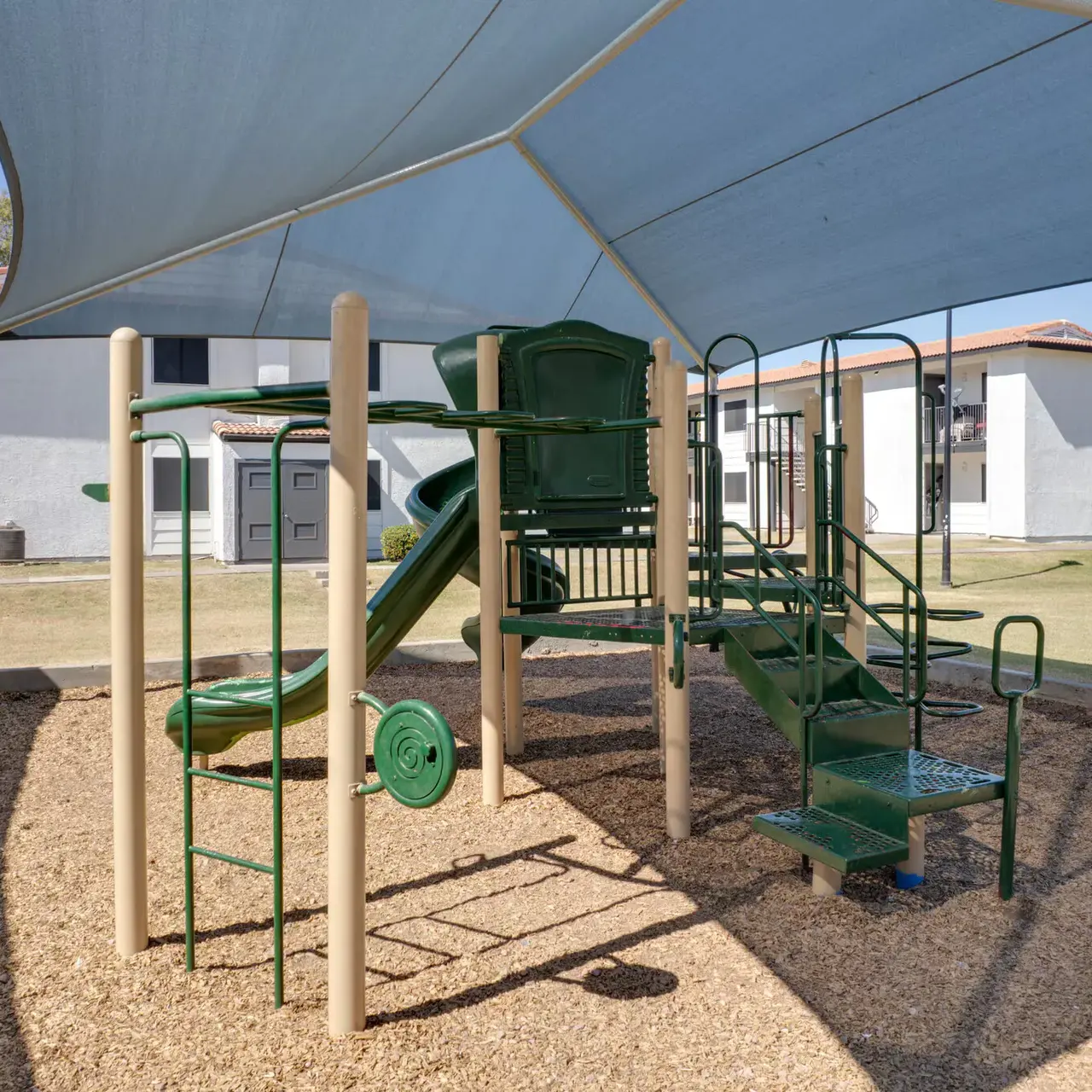 A green playground structure with slides and climbing features is covered by a blue shade. The surrounding area is filled with wood chips, and there are two white buildings in the background.