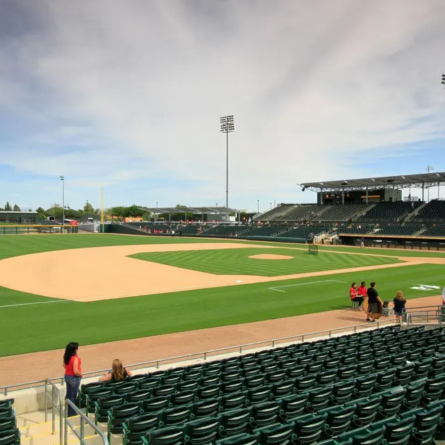 A panoramic view of a baseball diamond with a well-maintained infield and outfield grass. Empty bleachers surround the field, with a bright sky above and stadium lights in the background.
