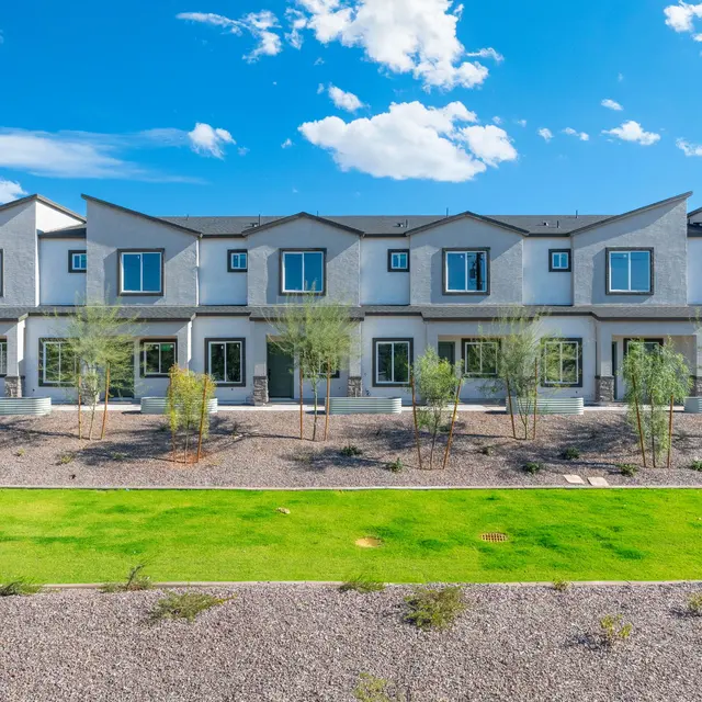 Modern Townhouses A row of modern townhouses with a clear blue sky and scattered clouds overhead. The houses feature a minimalist architectural design with multiple windows and vegetation in front. The foreground includes green grass and rocky ground.