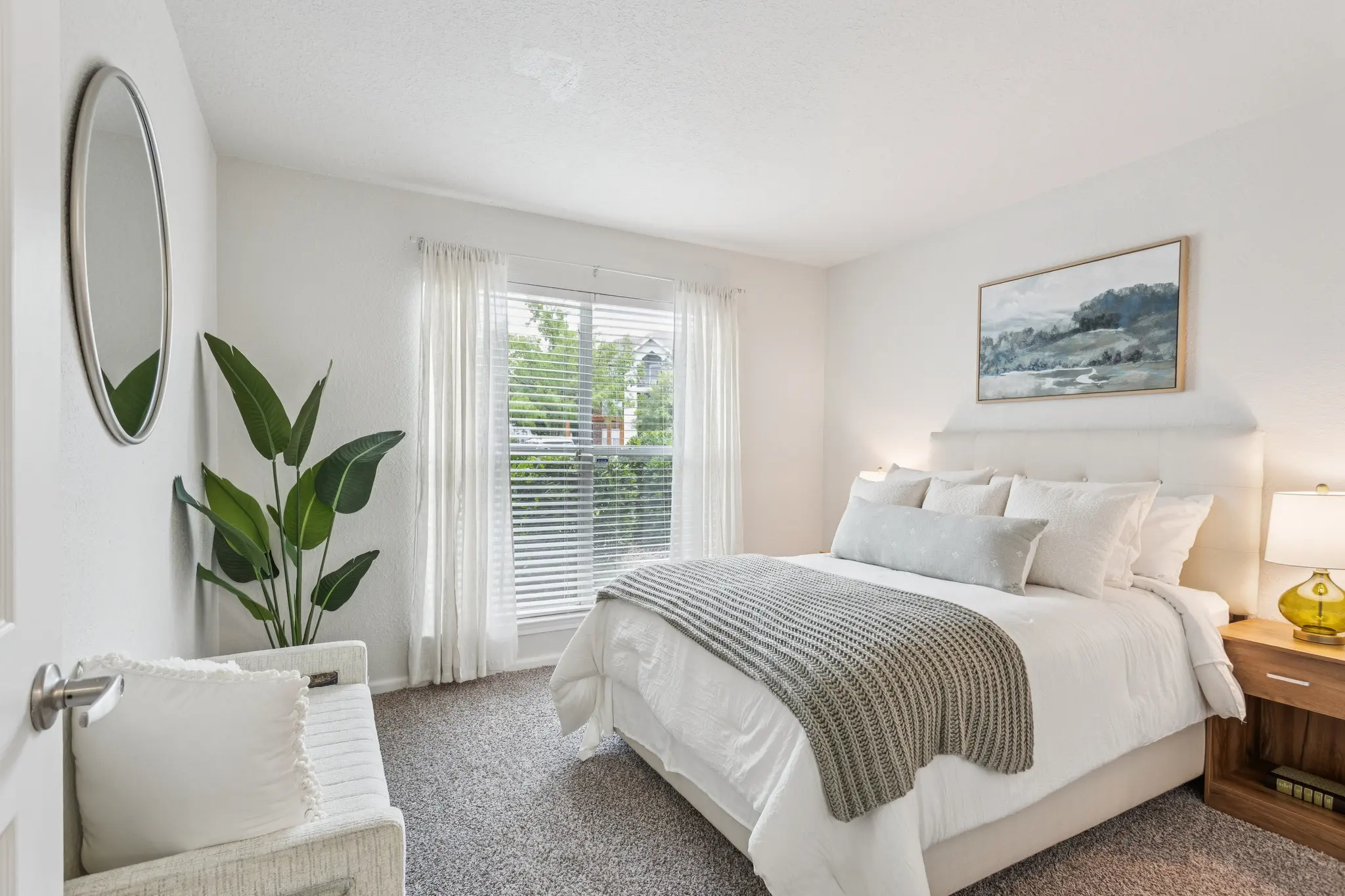 A modern bedroom featuring a large bed with white bedding and a grey throw blanket. There is a round mirror on the wall, a plant in the corner, and a window with sheer curtains allowing natural light to enter.