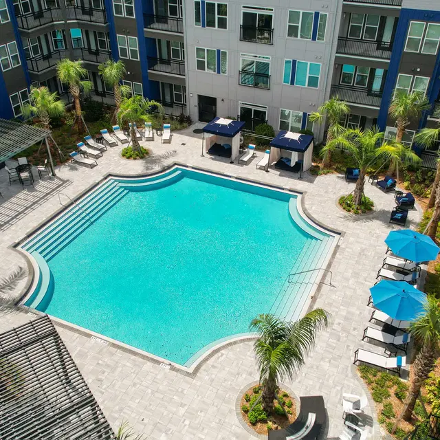 Aerial view of a modern apartment complex featuring a large swimming pool surrounded by palm trees and lounge chairs.