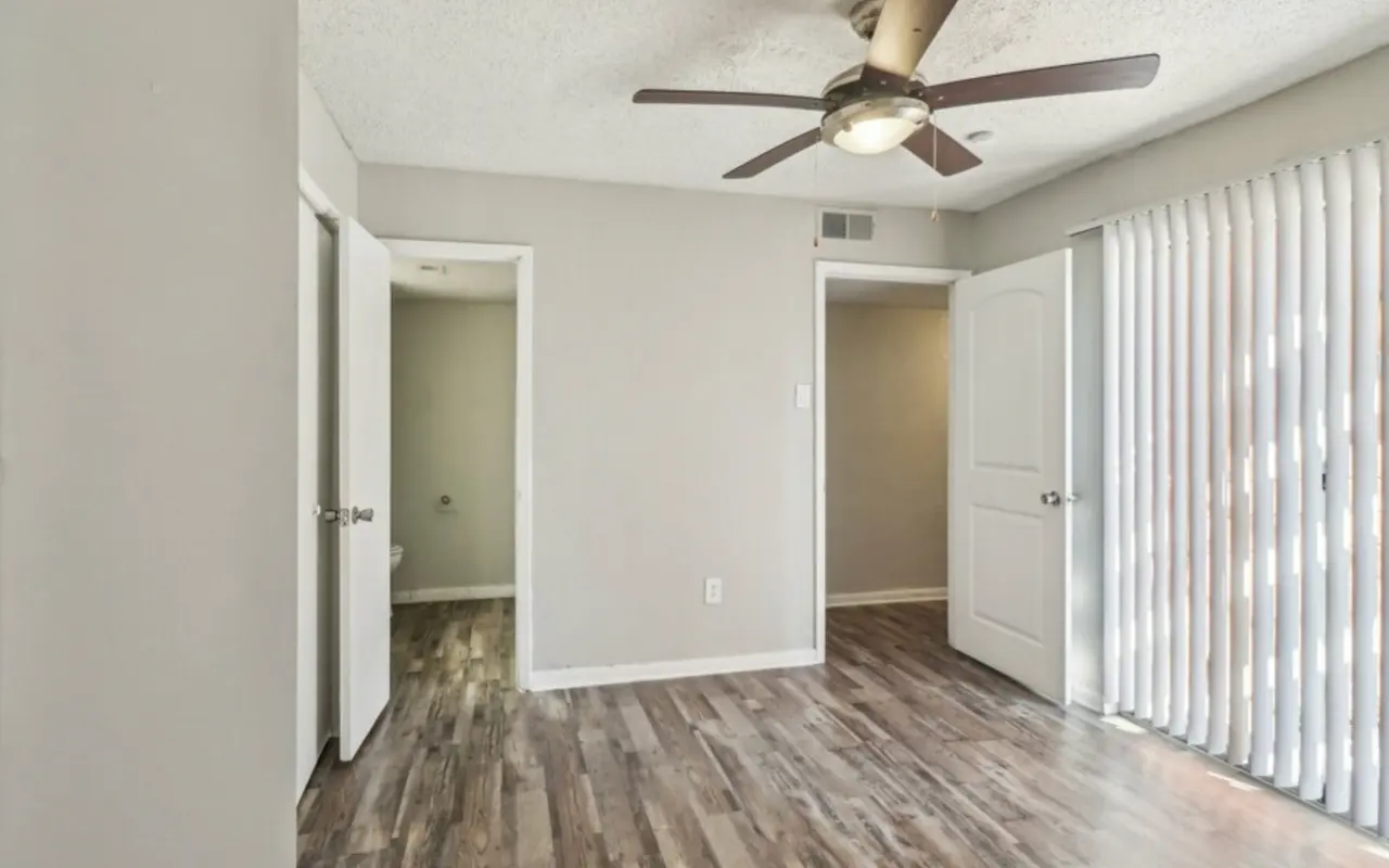 A well-lit interior room showcasing a ceiling fan and wooden flooring, with two doors visible that lead to separate areas. One door is slightly ajar, and the room features vertical blinds on a window.