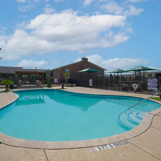 A clear blue swimming pool surrounded by a concrete deck, with patio seating and umbrellas in the background under a blue sky.