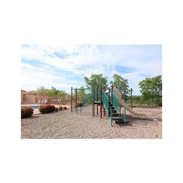 Playground with Slides A playground featuring green slides and climbing structures on sandy ground, surrounded by shrubs and trees under a blue sky with scattered clouds.