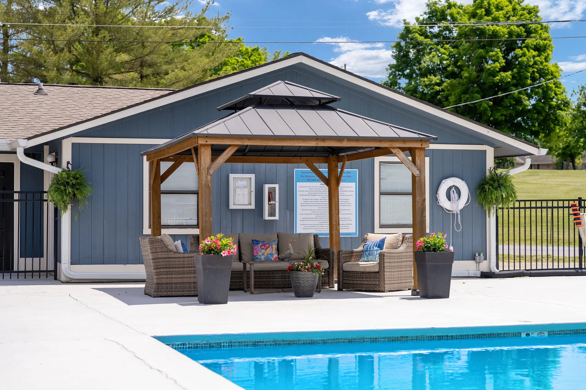 A cozy gazebo with seating next to a swimming pool, surrounded by greenery and bright blue sky.