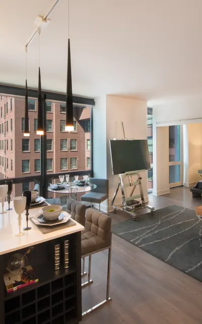 A contemporary apartment interior featuring a living area with large windows showcasing a city view, a dining table set for two, modern furniture including a tan couch, a stylish coffee table, and an open kitchen area with bar stools.
