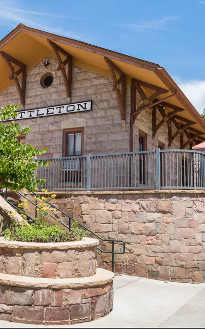A historical stone train station building with a steep roof and decorative wooden brackets. The building is surrounded by trees and shrubs, with a staircase leading up to it.