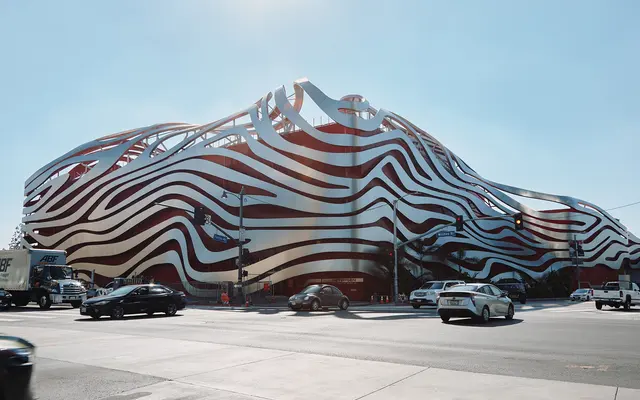 A modern, wavy architectural structure featuring a striped exterior in red and white. Located at a street corner, vehicles are seen passing by in the foreground, and the sky is clear blue.