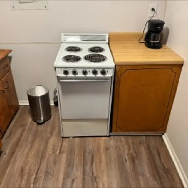 A corner of a kitchen featuring a white electric stove, a wooden countertop, a black coffee maker, and a trash can. The floor has a wooden appearance.