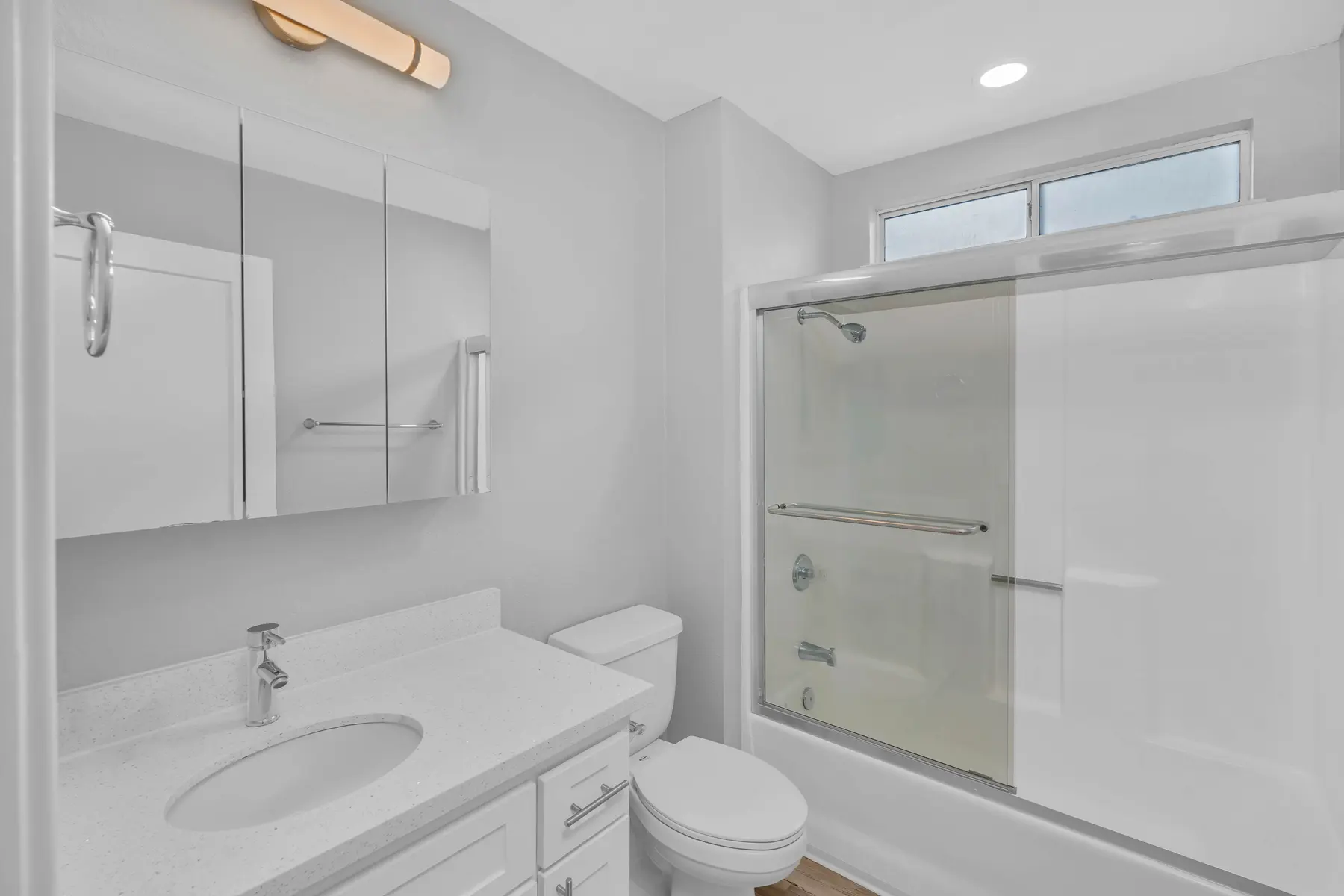 A bright and clean bathroom featuring a white vanity with a sink, a round mirror, a toilet, and a clear glass shower enclosure. Natural light comes in from a window.