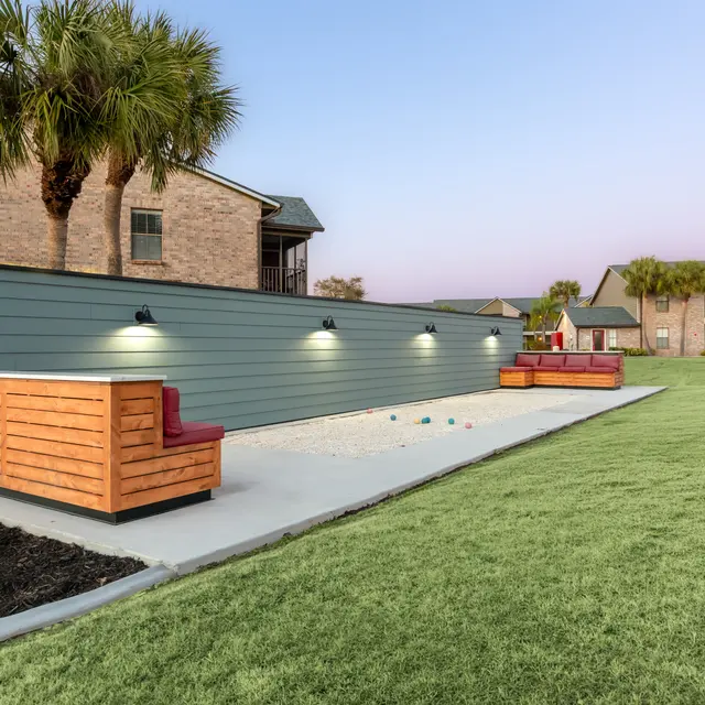 An outdoor lounge area featuring a long wall with lights, two wooden seating structures in different styles, surrounded by a lawn and palm trees, with nearby buildings visible.