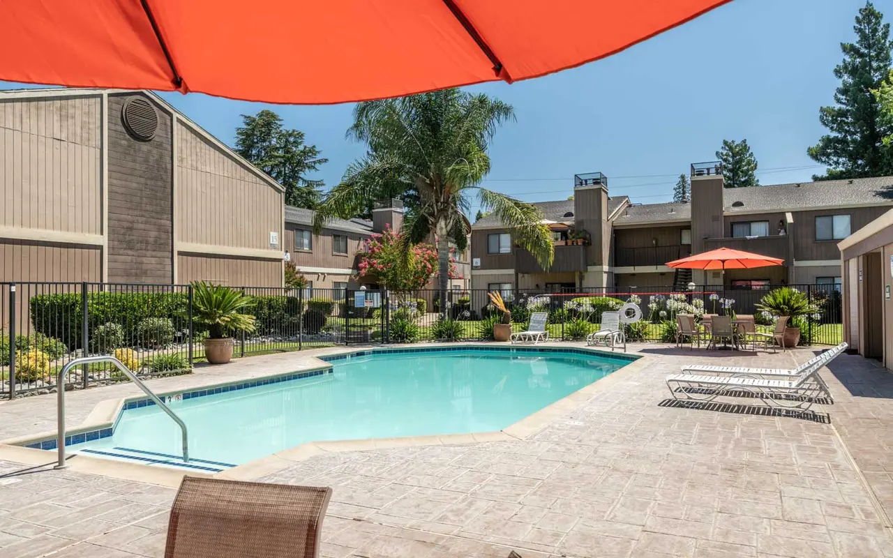 A well-maintained swimming pool area surrounded by lounge chairs and umbrellas, with buildings in the background and lush greenery.