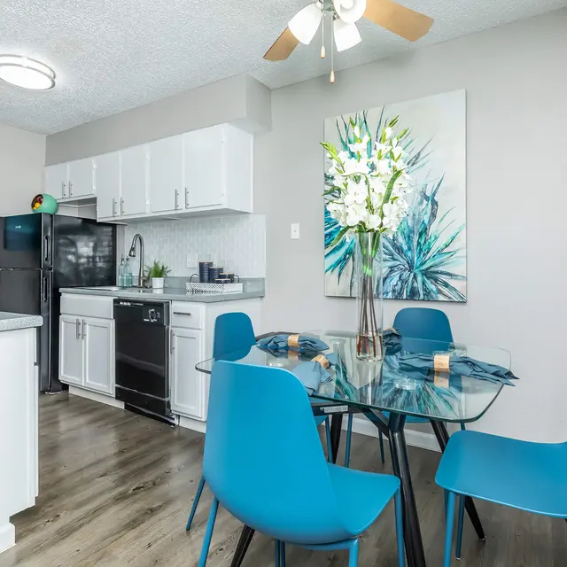 A modern kitchen and dining area featuring a glass dining table with blue chairs, white cabinetry, and vibrant wall art.