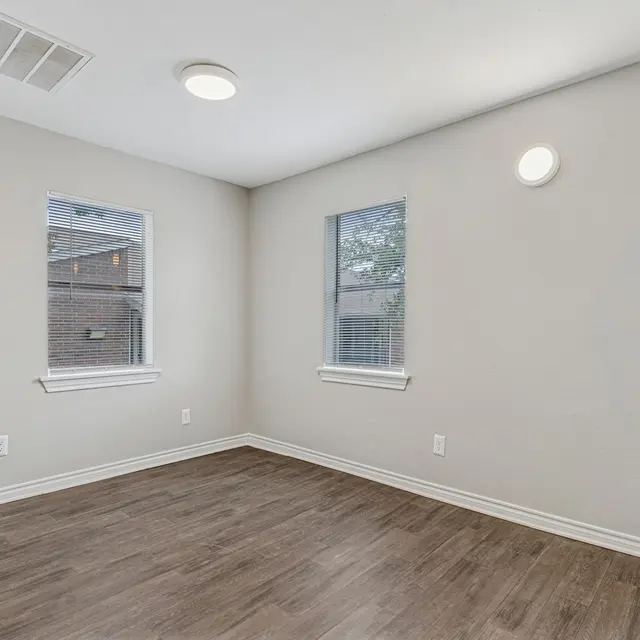 An empty room with light brown wooden flooring and two windows. The walls are painted light gray, and there is a circular ceiling light in the center. One window is partially open.