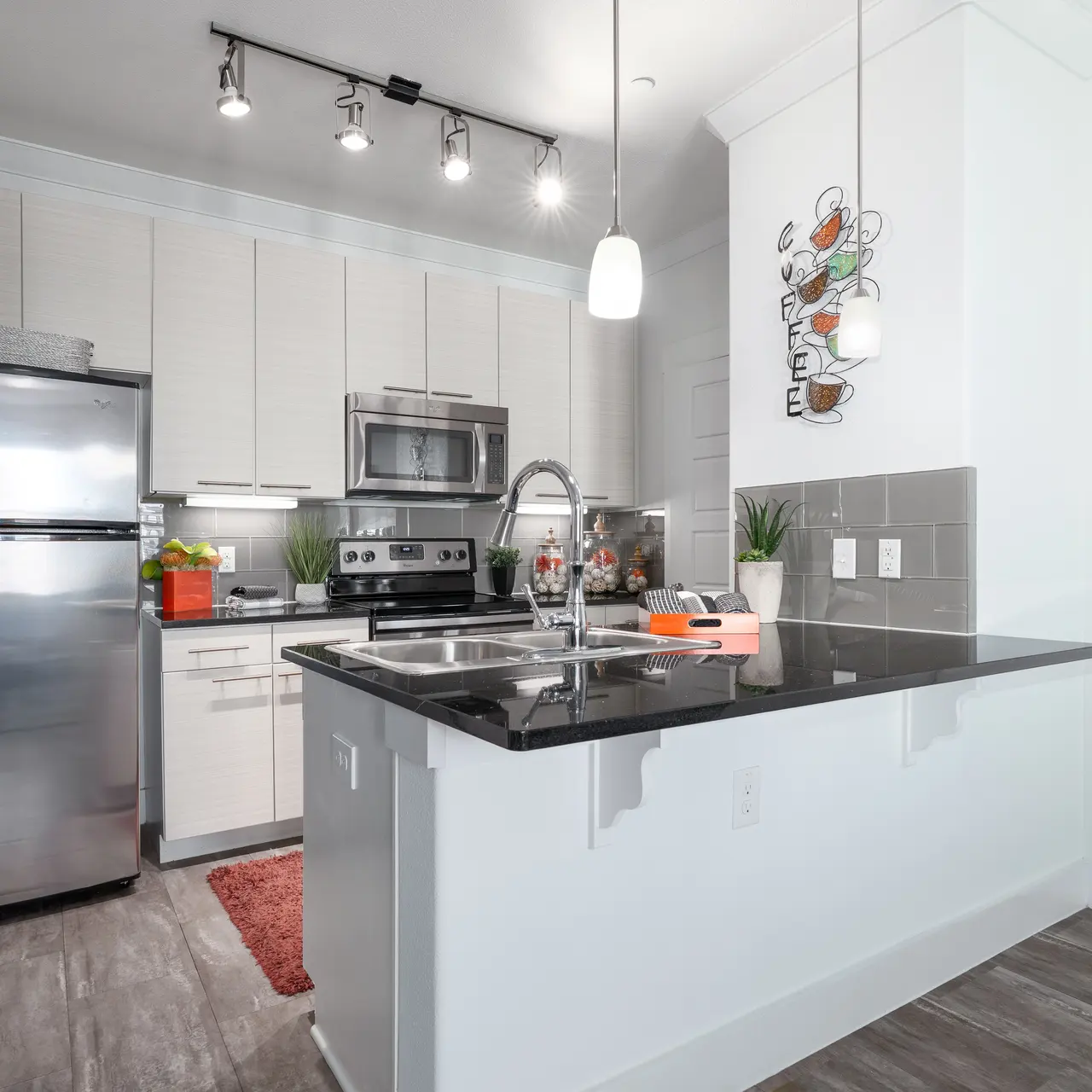 A modern kitchen featuring stainless steel appliances, a black countertop, and contemporary cabinets. The kitchen island has a small sink and is complemented by bright lighting and decorative elements on the wall.