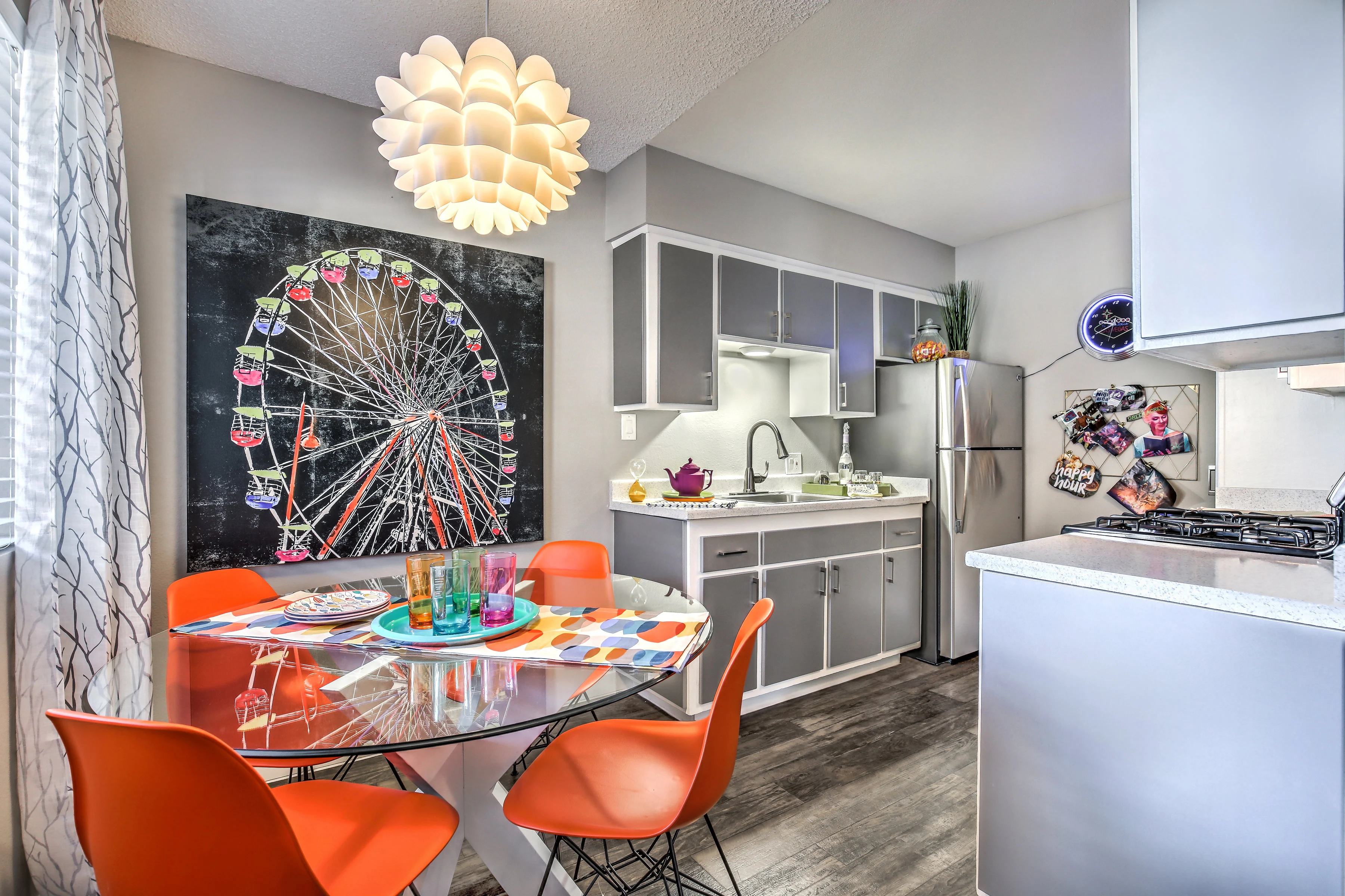 Stylish Apartment Kitchen and Dining Space A modern kitchen and dining area featuring a round glass table with colorful chairs, a large artwork of a ferris wheel on the wall, and stainless steel appliances in the kitchen.