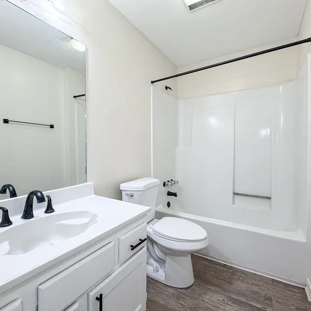 A modern bathroom featuring a white bathtub and shower combo, a white vanity with black faucet, and a mirror above the sink. The walls are painted light colors, and there is a wooden floor.