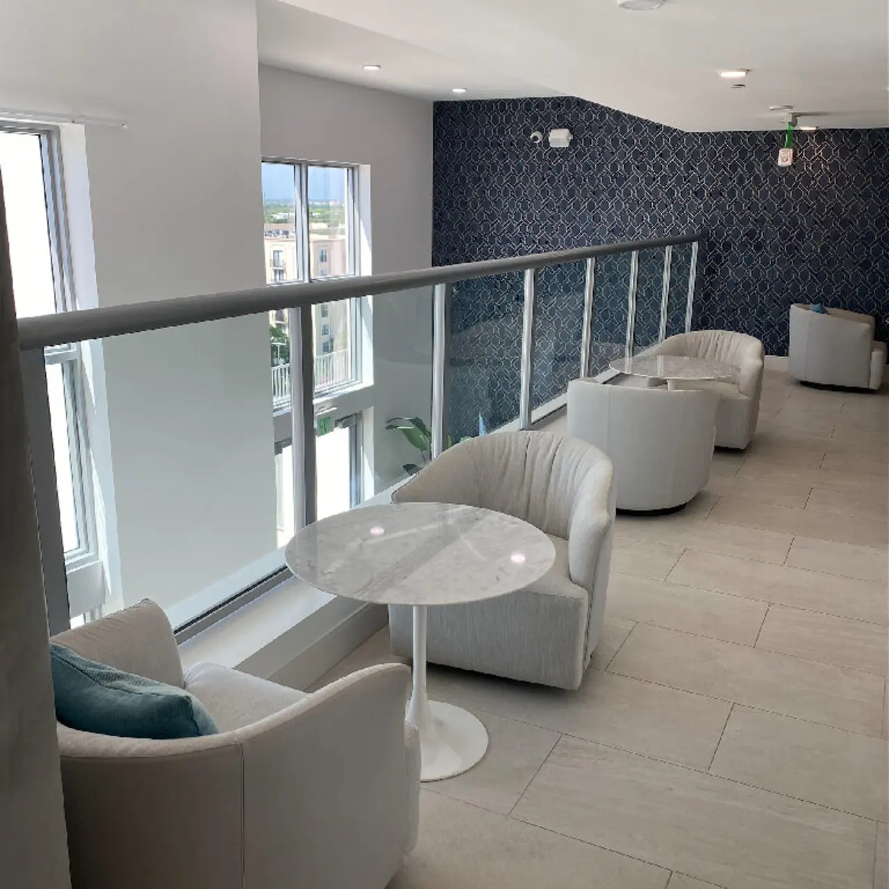 A modern lounge area features white chairs and a round table, with large windows allowing natural light, and a patterned wall in the background.
