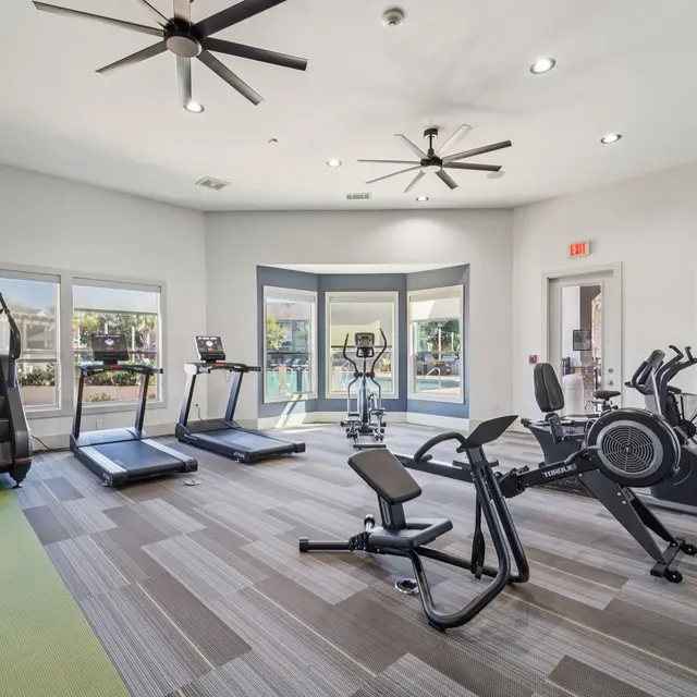 Interior view of a modern fitness center with exercise equipment including treadmills, stationary bikes, and elliptical machines, with large windows allowing natural light.