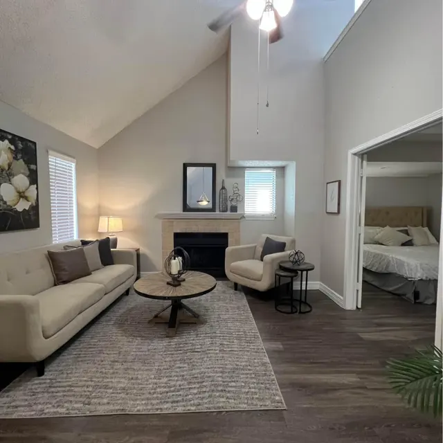 A modern living room with a high ceiling, featuring two beige sofas, a round coffee table, and a cozy fireplace. Natural light filters in through the windows, and there is a view of a bedroom through an open doorway.